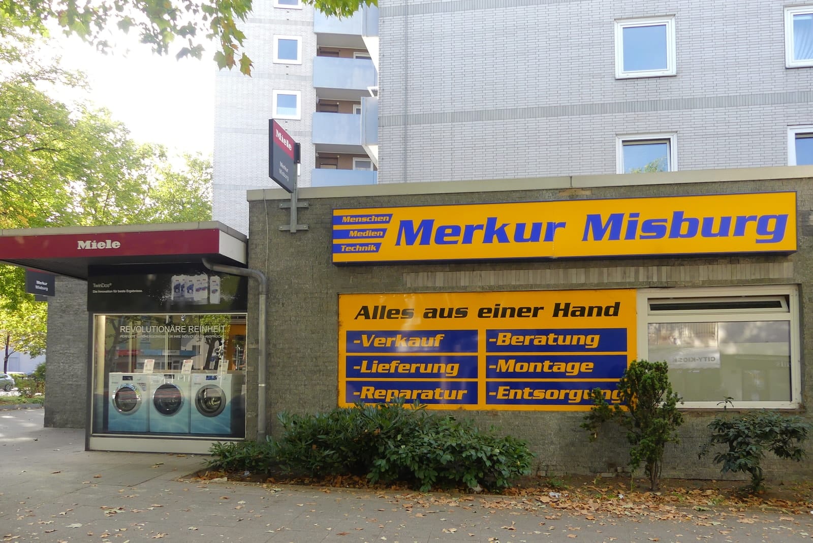 Gebäudeecke mit Merkur-Schild und der Tafel Alles aus einer Hand: Verkauf, Beratung, Lieferung, Montage, Reparatur, Entsorgung