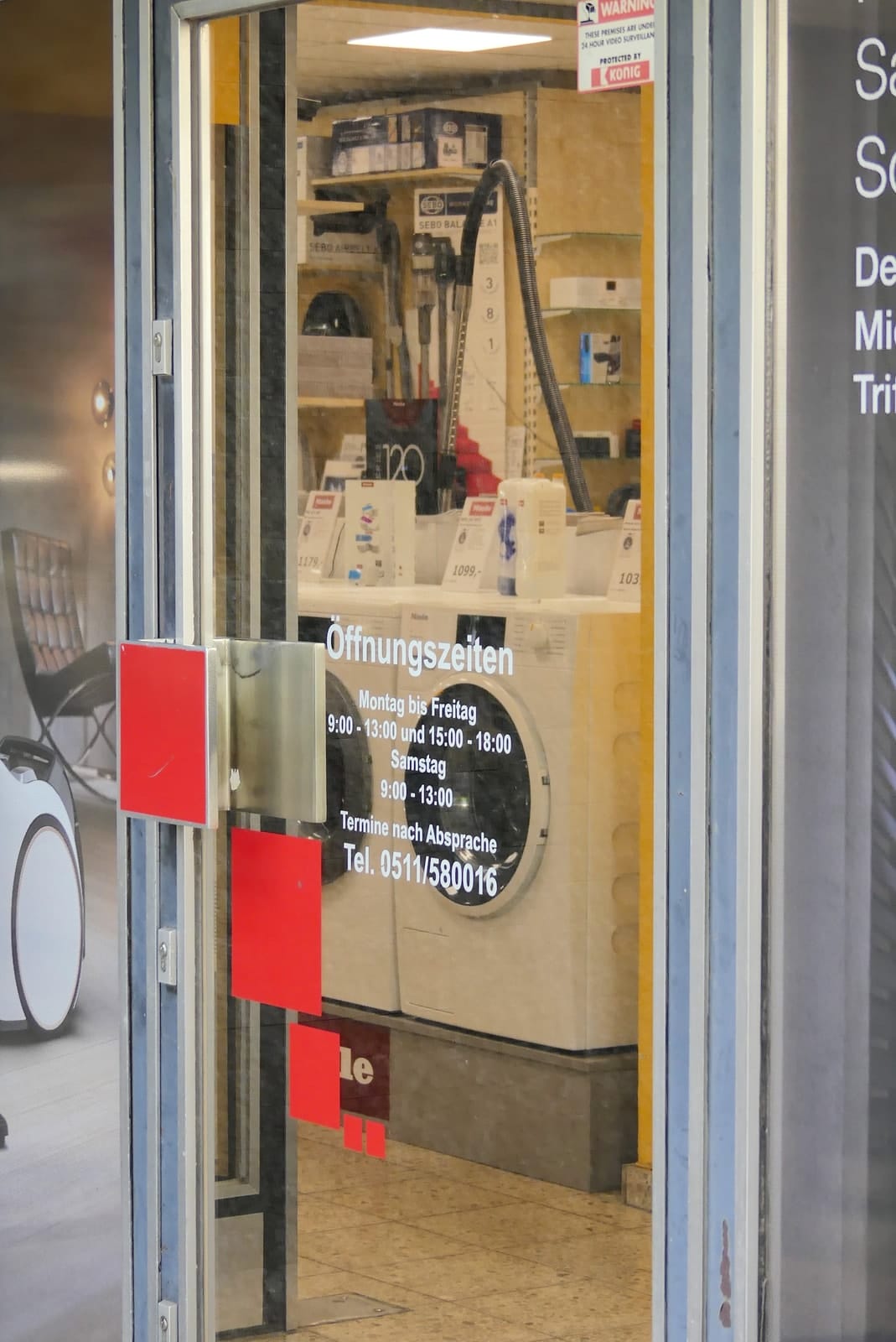 Glastür des Geschäfts mit aufgeklebten Öffnungszeiten und Telefonnummer, dahinter Staubsauger in der Ausstellung