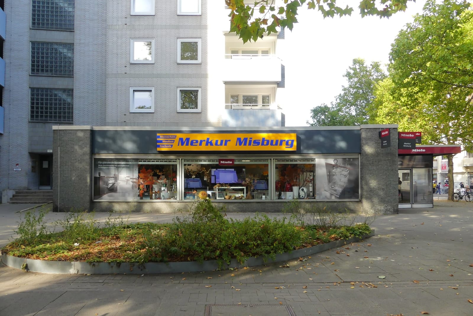 Das Geschäft Merkur Misburg mit Vorplatz und bepflanztem Beet an der Waldstraße