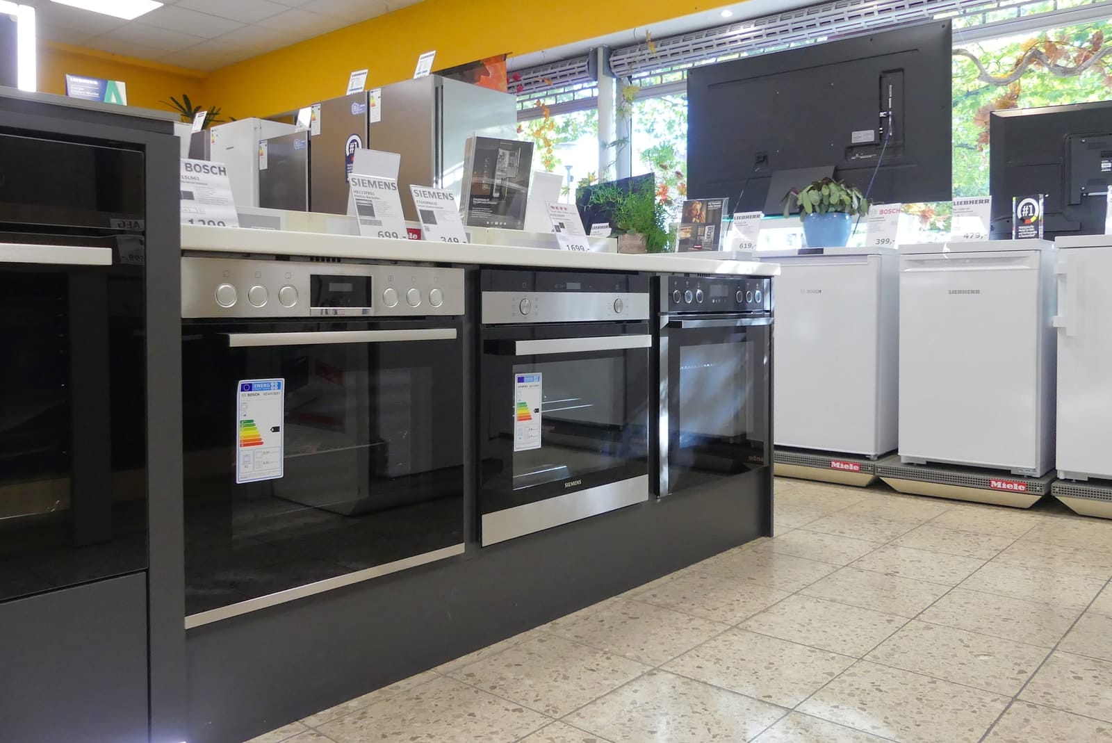 Reihe von Einbaubacköfen in dunkler Front, dahinter weiße Kühlgeräte und Fernseher