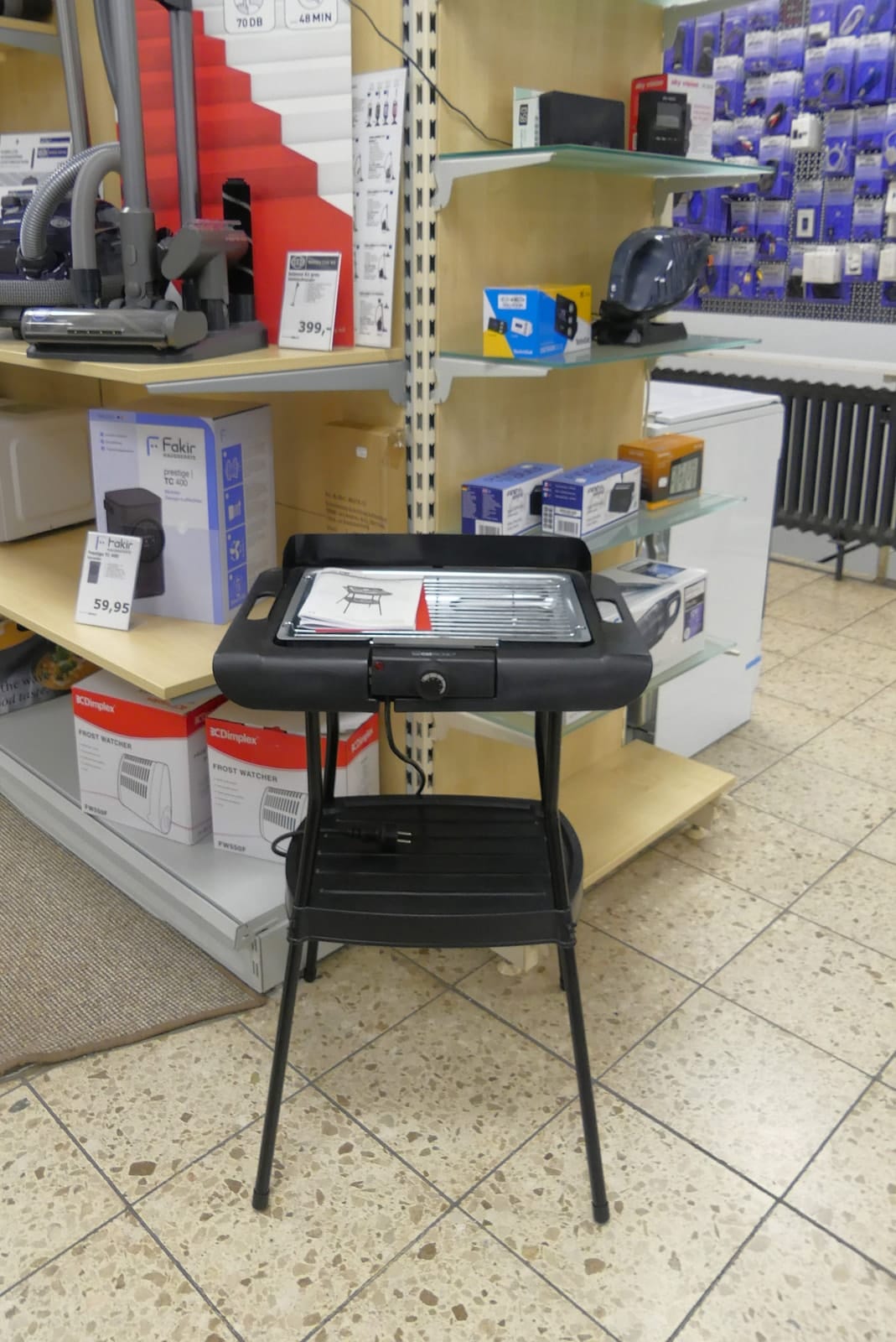 Elektrischer Standgrill auf schwarzem Gestell zwischen Regalen mit Staubsaugern und Zubehör