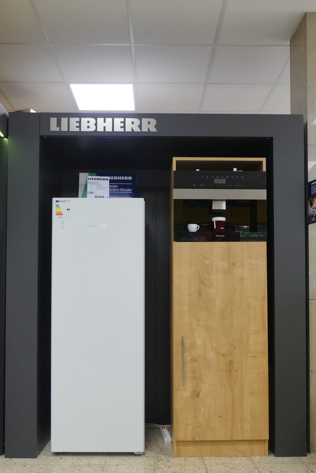 Liebherr-Präsentation mit weißem Standkühlschrank und Einbaugerät hinter Holzfront