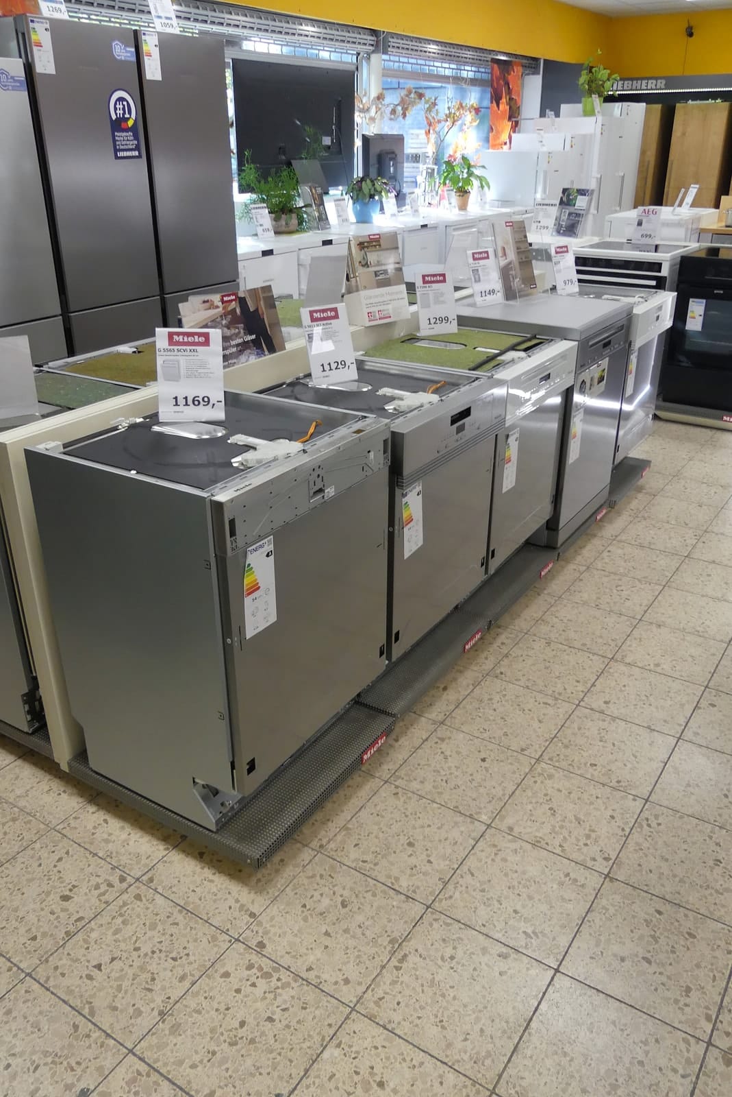 Reihe von Geschirrspülern mit Edelstahlfront und Preisschildern in der Ausstellung, dahinter Kühlschränke